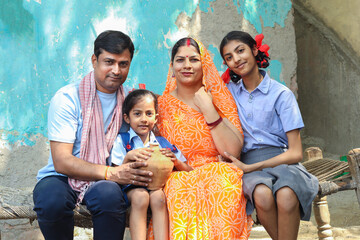 Rural indian family sitting with gullak or piggy bank to save money. Parents with two daughter in school uniform looking at camera. Togetherness. People of india. Education of girls.