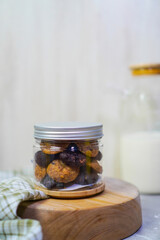 a jar filled with cookies placed on a wooden plate partially covered by gingham fabric, with milk bottles in the background