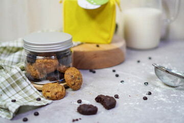 a jar filled with cookies and surrounded by chocolate chips, yellow bags with white and green tags placed on a wooden pedestal behind it, and milk bottles in the background