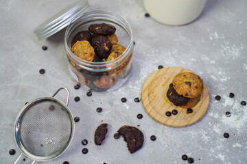 top view of an open jar filled with cookies, and some cookies outside the jar, surrounded by chocolate chips with flour sieve and the cropped milk bottle in the image