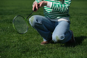 A person sitting on the grass with kneeled jeans, holding a badminton racket. The sunny weather...