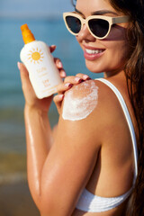A beautiful young woman applies sunscreen to her shoulder while enjoying a sunny day at the beach....