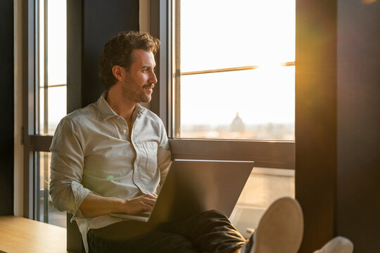 Man in office with laptop at sunset looking contemplative