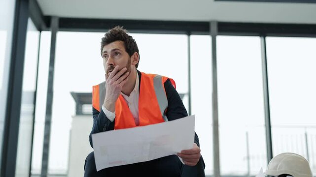 Frustrated architect sits on the floor in modern office building with blueprints. He's wearing suit and orange vest. He's unsure about something.