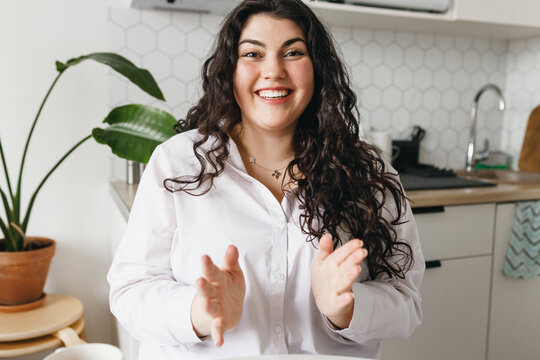 Portrait of excited young caucasian plus size woman gesticulating while talking, sitting at kitchen table, sharing funny story having video call or recording podcast for social media