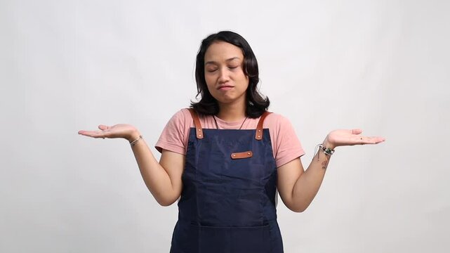 Young female barista wearing a navy apron making a confused expression, raising both hands in a shrug gesture. Studio shot with white background, representing doubt, uncertainty, or not knowing
