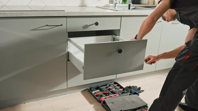 Man assembling kitchen drawer in modern cabinet using screwdriver. Handyman installing and adjusting drawer slide mechanism in contemporary kitchen. Furniture installation