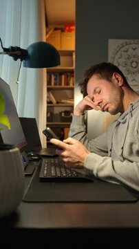 Tired freelancer procrastinating at home office, scrolling phone instead of working. Unproductive man using smartphone at home desk. Concept of procrastination and distraction in remote work