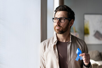 Man holding blue ribbon supporting prostate cancer awareness, looking out the window