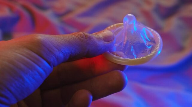 Hand Holding Condom Close-Up. Safe Sex Concept with Condom. In the Background, the Bed is Illuminated with Colored Intimate Evening Light