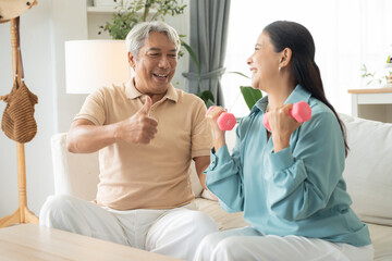 Obraz premium Asian senior couple exercise with lifting dumbbell for strength training on couch together at home. Husband showing thumbs up to support his wife. Healthy lifestyle people insurance concept.