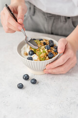 Bowl of granola with greek yogurt, kiwi and blueberries and spoon in hands of woman on white table.