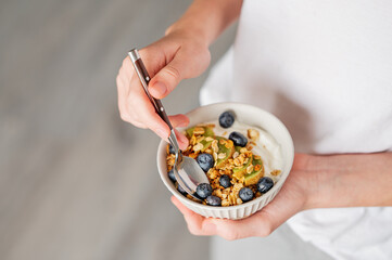 Bowl of granola with greek yogurt, kiwi and blueberries and spoon in hands of woman