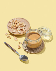 Peanut butter in glass jar on yellow background with spoon and salted nuts.