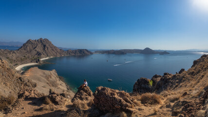 A breathtaking panoramic view showcases a man sitting on a rock, serene coastline featuring crystalclear waters alongside rugged rocky terrain of Padar Island all beneath bright sky, Komodo, Indonesia