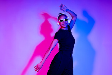 Person dancing in a studio with neon lights, wearing wireless headphones and sunglasses.