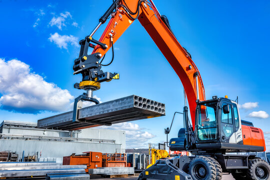 Orange mobile excavator lifting concrete structural elements at industrial worksite. Heavy machinery with hydraulic arm and grabber attachment handling precast materials. Construction equipment