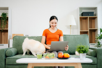 Woman relaxing on sofa at home with dog reading digital tablet in bright modern living room, joyful pet