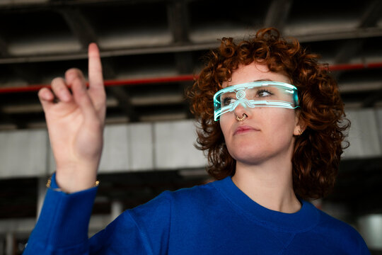 Young woman with curly hair wearing VR glasses and pointing indoors