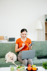 Young woman sitting on white sofa and working indoors holding small white puppy in bright modern living room, joyful pet