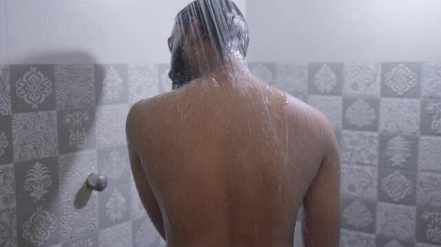 A back shot of a South Asian man rubbing his body with hot water in the shower under bright natural light
