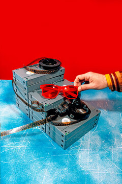 Hand holding red sunglasses over stacked VHS cassettes on a blue table with a red background