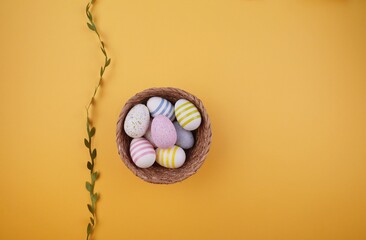 A wicker basket filled with colorful Easter eggs sits on an yellow background, accented by a green...