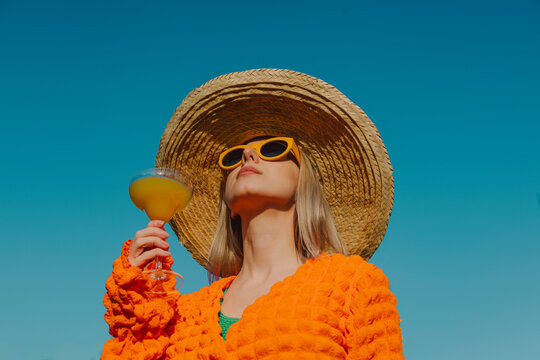 Stylish woman in orange clothing and straw hat enjoying a cocktail outdoors