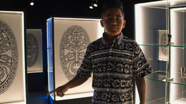 Young maori boy, dressed in a traditional patterned shirt, stands in a museum exhibition, gesturing towards maori artwork and cultural symbols, proudly sharing his heritage