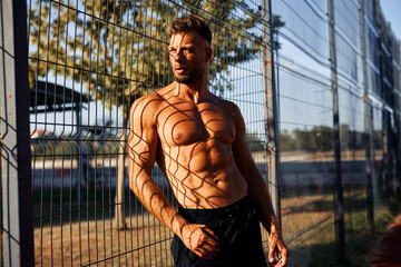 Muscular man with a defined six-pack leaning against a wire mesh fence, casting interesting shadows...