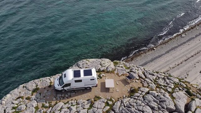White camper van with solar panels parked on a rocky cliff overlooking a beautiful turquoise ocean, offering a serene and idyllic camping spot