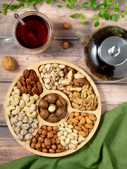 A high-angle flat lay shot showcasing a healthy assortment of nuts and a hot beverage on a rustic, light brown wooden surface. The central element is a round wooden serving tray with six compartments,