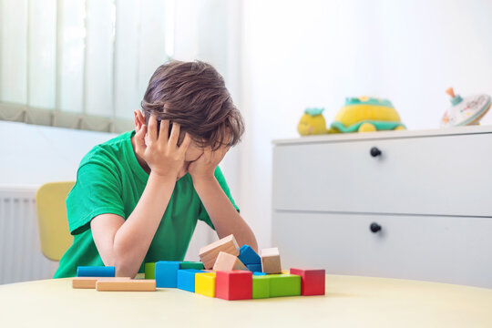 Tired little boy covering face with hands after playing with wooden blocks, experiencing frustration and emotional meltdown, focus on early child development, speech therapy, mental health support
