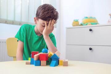 Tired little boy covering face with hands after playing with wooden blocks, experiencing...