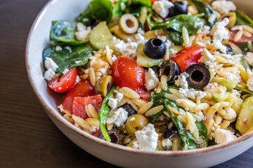 healthy orzo salad served in a bowl, made with fresh vegetables, black olives, and crumbled feta cheese close up