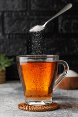 White sugar falling from teaspoon into glass cup of hot tea placed on coaster against black brick wall as background