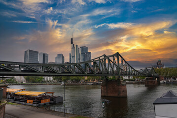 Eiserner Steg spans the Main River in Frankfurt, Germany, with a docked boat, historic cathedral spire, and modern skyscrapers under an overcast sky.