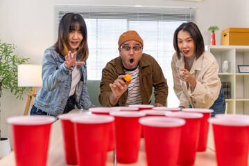 Happy asian diverse group friends playing with beer pong at home office standing at the table enjoying time together. Office leisure and board games concept.