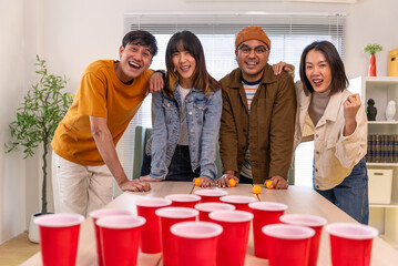Happy asian diverse group friends playing with beer pong at home office standing at the table enjoying time together. Office leisure and board games concept.