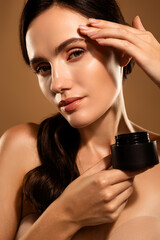 Young woman demonstrating skincare routine holding a black cosmetic jar on a pastel vertical...