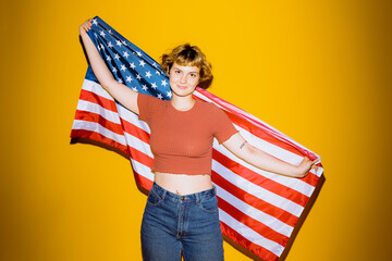 A young individual joyfully holds the American flag against a bright yellow backdrop. Their...