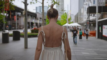 A woman with her blonde hair tied up in a bun walks down a busy city street wearing a summer white dress with an open back and shoulder ties.