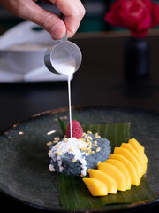 A plate of ripe mango with sticky rice and coconut milk sauce pouring on it, various sweet Thai desserts in background with Thai style plate decorations such as green banana leaves 