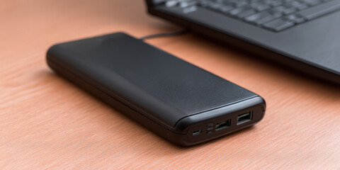 Black power bank with USB ports connected to a laptop on a wooden surface.  