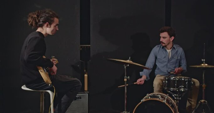 Two musicians, a bass guitarist and drummer, collaborate during a practice session in a dimly lit studio, preparing for an upcoming concert.