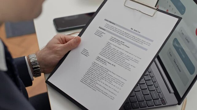 Cropped shot of male employee holding folder with CV of candidate when sitting in front of laptop at table during job interview at office