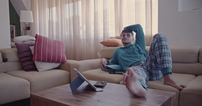 Young man relaxing on the sofa in his pajamas, enjoying his day off. He watches TV with a remote in his hand.