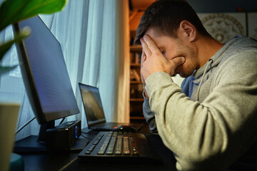Exhausted man sitting at computer with face in hands. Freelancer struggling with burnout while...