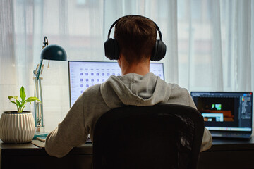 Man working at desk with two screens and headphones, back view. Focused freelancer editing digital...
