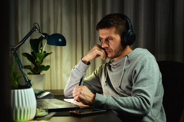 Focused man in headphones working on laptop at night and writing notes. Freelancer working remotely from home